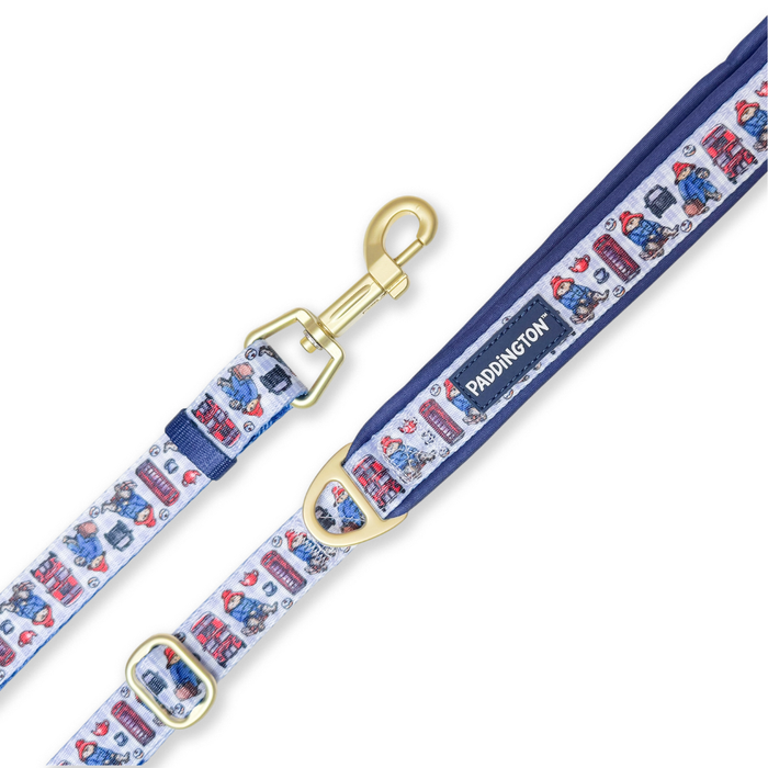 A Paddington-themed dog leash with a white background printed with red double-decker buses, telephone booths, and Paddington Bear illustrations, featuring a navy blue top handle and gold-tone metal hardware.