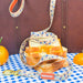 A patterned dog leash with a gold clasp and a orange Paddington brand tag, draped over a stack of bread slices with orange marmalade filling. The scene is styled on a blue-and-white checkered tablecloth with oranges and a brown leather suitcase in the background.