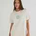A woman wearing an oversized off-white t-shirt with a green circular chest graphic that reads 'MATCH POINT PADDINGTON'.