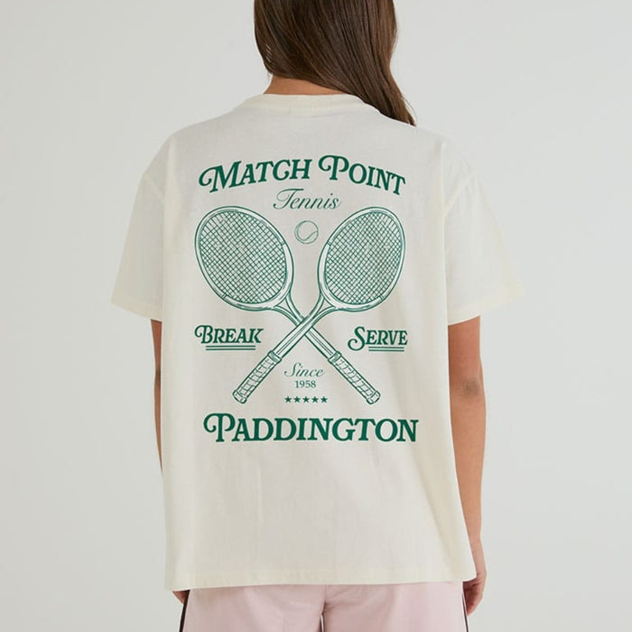 A woman viewed from the back wearing a cream-colored t-shirt with a green tennis-themed graphic print that reads 'MATCH POINT Tennis BREAK SERVE Since 1958 PADDINGTON' around two crossed tennis rackets.