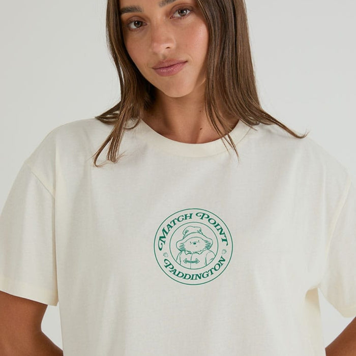 A woman wearing a cream-colored crewneck t-shirt with a green circular 'Match Point Paddington' logo printed on the chest, set against a plain light background.
