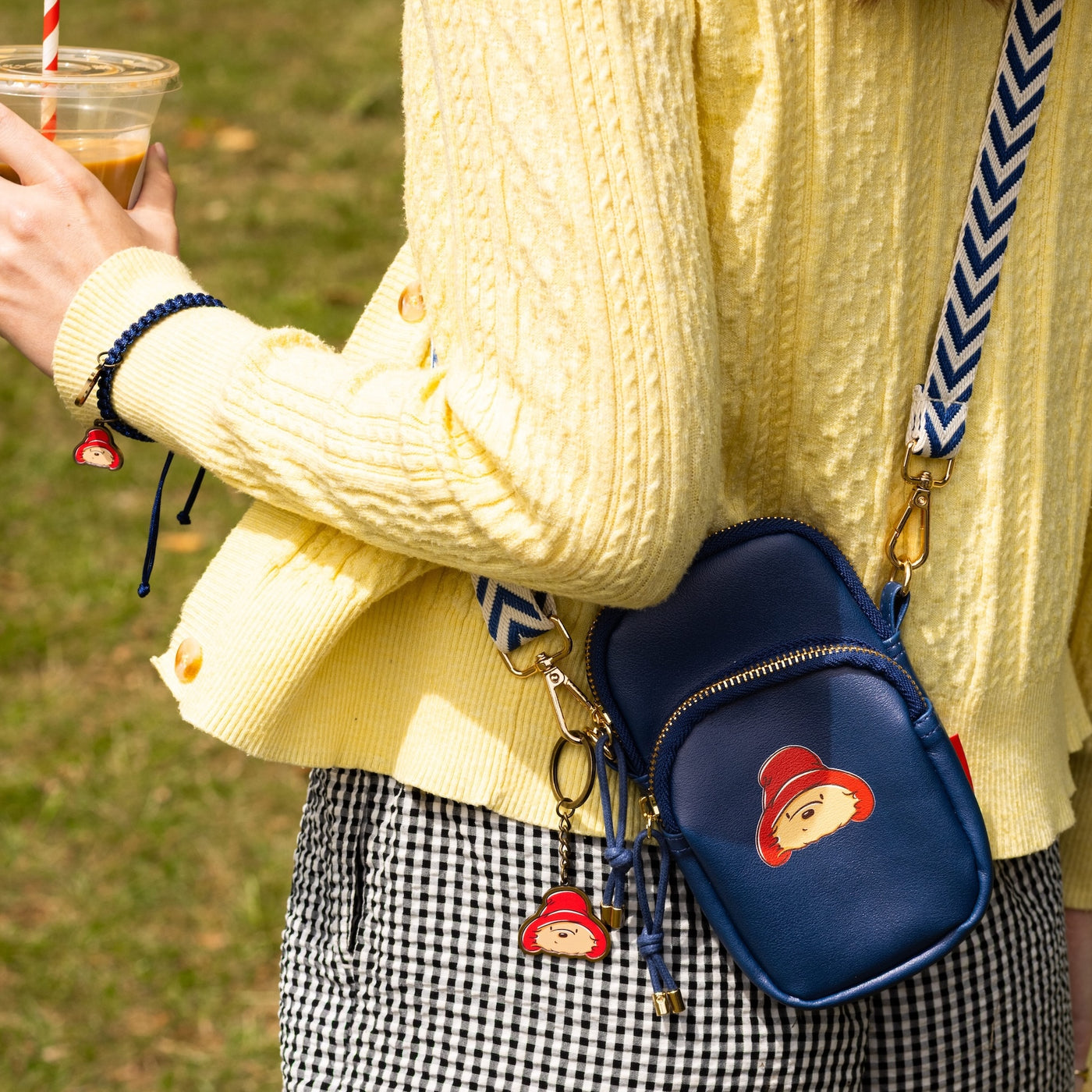 Shop The Look Paddington Essentials Collection - Person wearing a yellow sweater with a blue bag featuring a cartoon paddington, holding a drink.