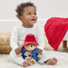 A baby sitting on a white rug holding a Paddington teddy bear plush toy dressed in a red hat and blue outfit, with a red textured throw blanket draped over a stool in the background.