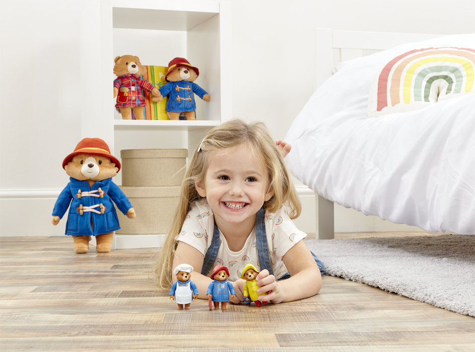 A little girl playing with Paddington bear toy figurines in a white bedroom and rainbow pillow