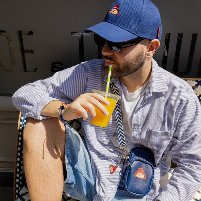 Man sitting outdoors drinking orange juice from a straw, wearing a navy blue Paddington cap and black sunglasses.