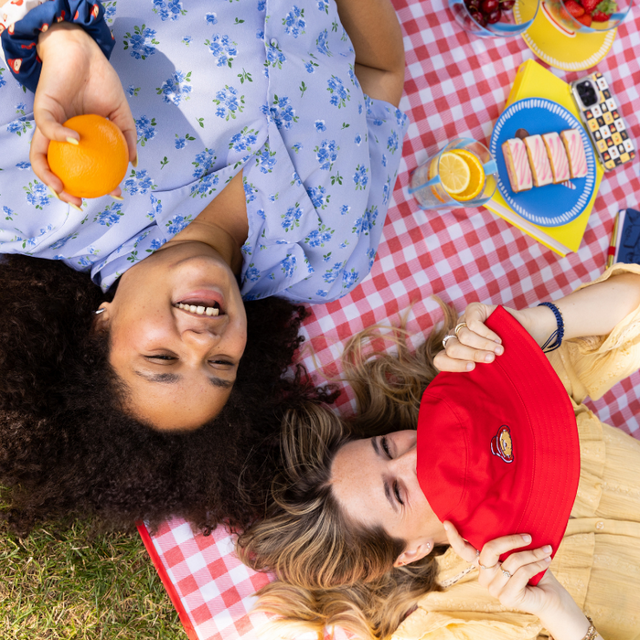 Two people lying on a checkered summer picnic blanket with food and drinks. One holding an orange and another holding a red paddington bucket hat