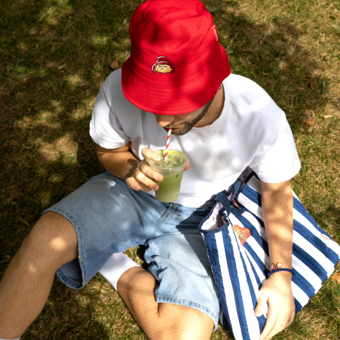 Person sitting on grass, wearing a red Paddington hat and white shirt, drinking from a green smoothie.