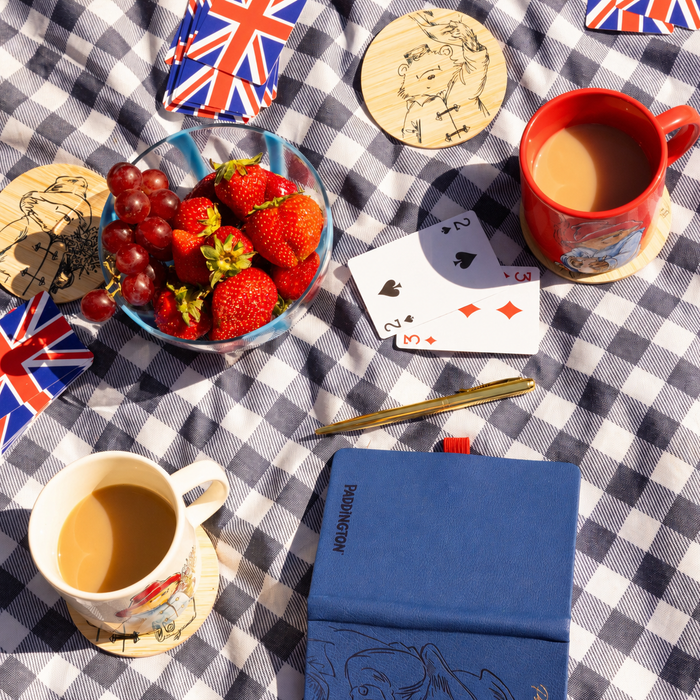 A styled picnic scene on a blue-and-white checkered blanket with a glass bowl of strawberries and red grapes, a red Paddington mug of tea, a white cup of tea, playing cards, a gold pen, a blue notebook, and small Union Jack playing cards.