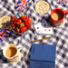 A styled picnic scene on a blue-and-white checkered blanket with a glass bowl of strawberries and red grapes, a red Paddington mug of tea, a white cup of tea, playing cards, a gold pen, a blue notebook, and small Union Jack playing cards.