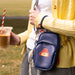 Person holding a blue Paddington pouch with a cartoon head logo, wearing a navy blue charm bracelet, a yellow sweater and plaid pants, outdoors.