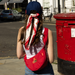 Person with a red paddington bag and scarf standing next to a red postbox on a street.