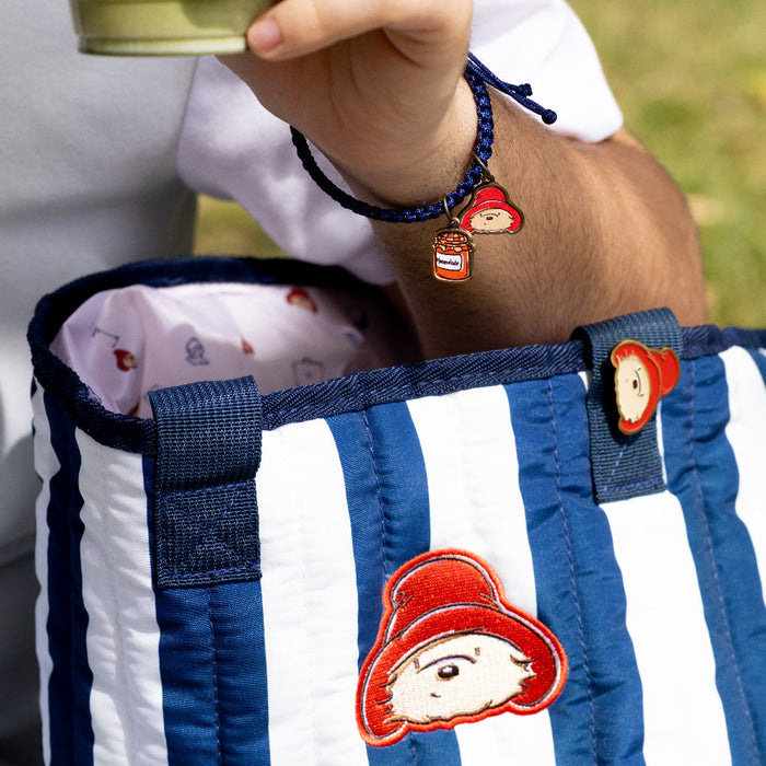 Blue and white striped bag with cartoon character patches, a paddington enamel pin badge and a charm bracelet, held by a person.