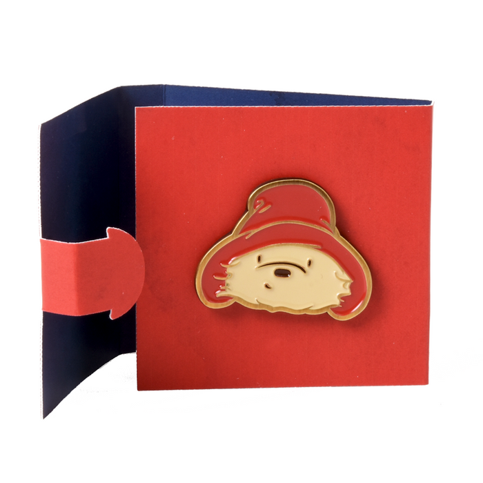 Red card envelope with a gold brass pin badge featuring a cartoon Paddington bear character on a white background