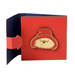 Red card envelope with a gold brass pin badge featuring a cartoon Paddington bear character on a white background