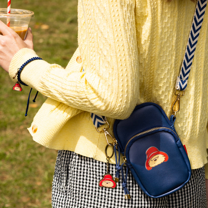 Person wearing a yellow sweater with a blue bag featuring Paddington bear and matching Paddington keyring, holding a drink.