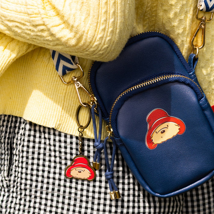 A navy blue faux-leather crossbody phone bag with a Paddington bear character patch and matching keyring charm, styled on a yellow knit sweater and black-and-white checkered skirt.