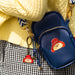 A navy blue faux-leather crossbody phone bag with a Paddington bear character patch and matching keyring charm, styled on a yellow knit sweater and black-and-white checkered skirt.