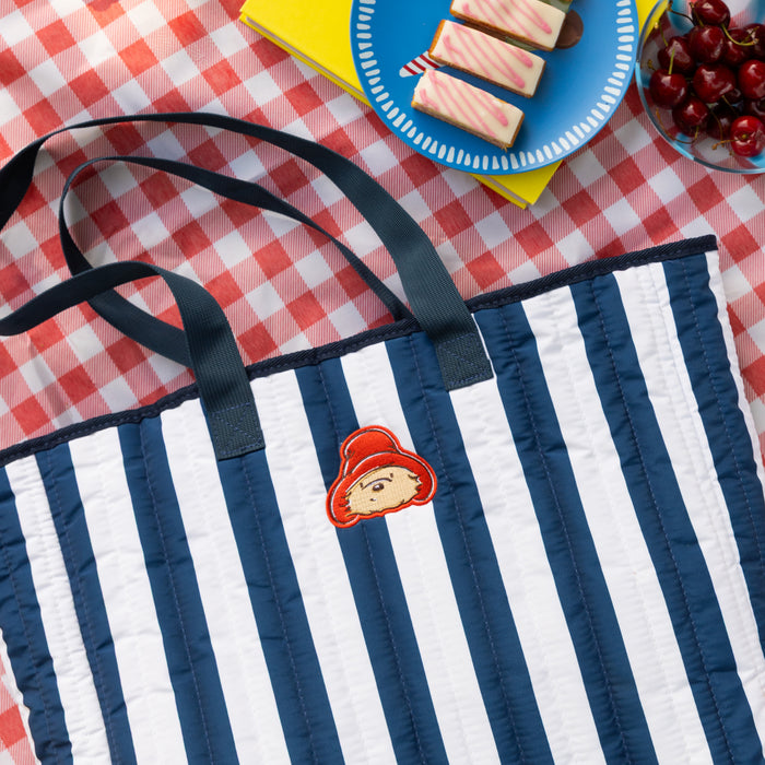 blue and white Striped tote bag with a cartoon Paddington bear character on a red and white checkered tablecloth with picnic items.