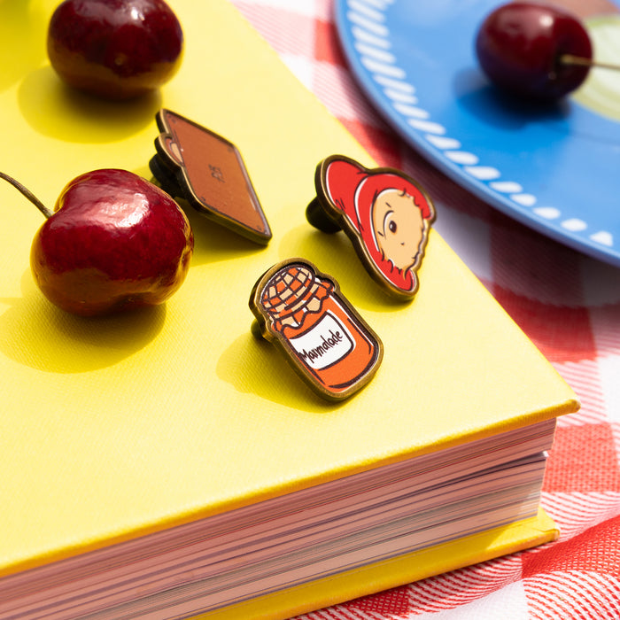 Three enamel Paddngton bear pins on a yellow book with cherries and a blue plate in the background.