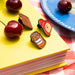 Three enamel Paddngton bear pins on a yellow book with cherries and a blue plate in the background.