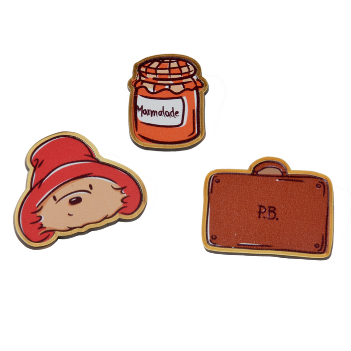 Three decorative pins featuring Paddington bear face logo, a jar of marmalade, and a brown suitcase on a white background.