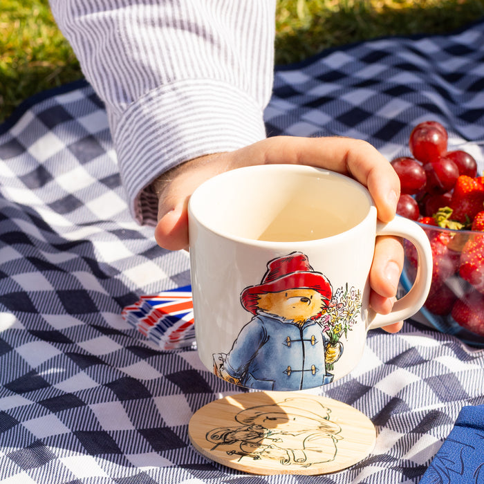 Person putting a mug with a Paddington Bear design on a Paddington bear coaster on a checkered blanket outdoors.