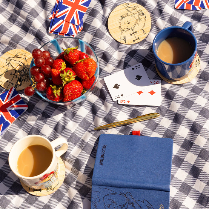 Table setting with coffee cups on Paddington coasters, strawberries, grapes, playing cards, and a blue notebook on a checkered tablecloth.