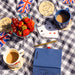 Table setting with coffee cups on Paddington coasters, strawberries, grapes, playing cards, and a blue notebook on a checkered tablecloth.