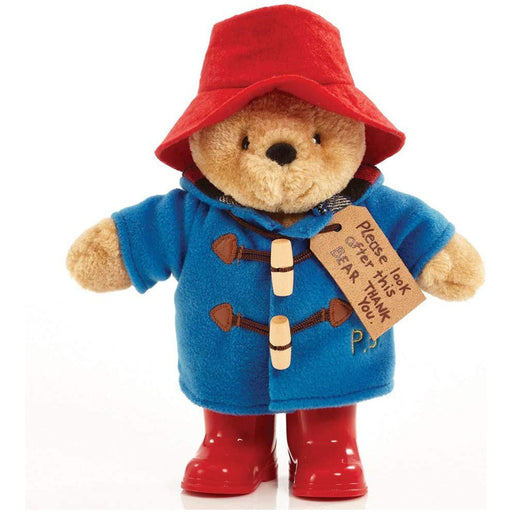 A plush toy of Paddington Bear wearing a blue duffle coat, red hat, and red boots, with a 'Please look after this bear. Thank you' tag around its neck.