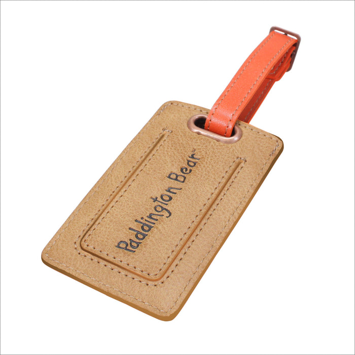 Brown leather luggage tag with 'Paddington Bear' text and orange strap on a white background